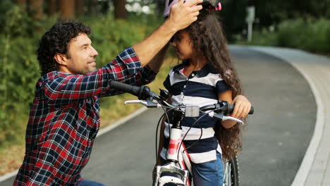 Closeup.-Portrait-of-a-pretty-girl-and-her-father-near-the-bike.-Dad-wears-a-helmet-on-the-girl's-head.-They-look-at-each-other.-Smiling.-Blurred-background